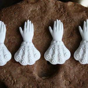 May include: Four white ceramic figurines of hands with intricate detailing, resembling a pair of hands clasped in prayer. The figurines are set against a brown, textured background.