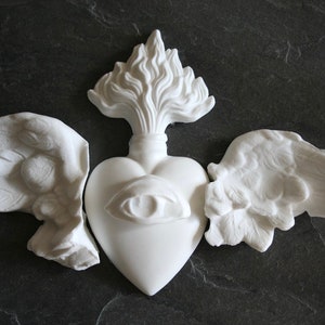 May include: White plaster sculpture of a heart with wings and a flame on top. The heart has an eye on the front.