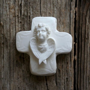 May include: A white, cross-shaped religious ornament with a sculpted angel figure at the center. The angel has detailed wings and curly hair. The cross is set against a weathered, wooden background.