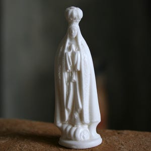Può includere: Una piccola statua bianca di una figura religiosa, forse la Vergine Maria, con una corona e vesti drappeggiate. La statua è in piedi su una base rotonda. La figura è in piedi con le mani giunte in preghiera.