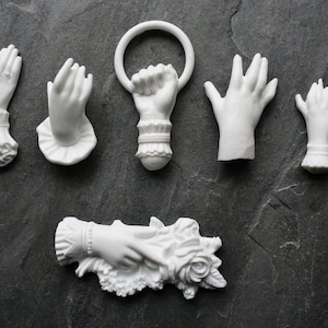 Könnte beinhalten: Satz von sieben weißen Porzellan-Handfiguren. Die Hände sind in verschiedenen Posen, darunter das Halten eines Rings, eines Blumenstraußes und eines Herzens. Die Figuren sind mit zarter Spitze und floralen Akzenten verziert.