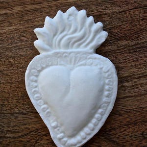 May include: A white, heart-shaped religious emblem with a flame design at the top. The heart has a detailed border and a textured surface. The emblem is set against a dark wood grain background.