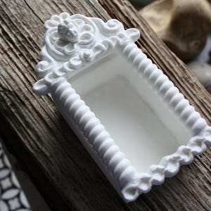 Peut inclure: Une étagère murale rectangulaire blanche et ornée, avec un motif floral et enroulé. L'étagère a une bordure en forme de corde et un bord festonné. L'étagère est faite d'un matériau solide.