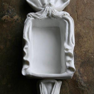 May include: White ceramic trinket dish with a decorative scalloped top and a rectangular center. The dish is shaped like a stylized flower.