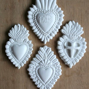 Puede incluir: Cuatro adornos de cerámica blanca en forma de corazón con diseños intrincados. Cada corazón presenta un diseño diferente, incluyendo una cruz, un corazón con las letras "GR" y dos corazones lisos.