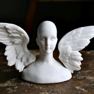 May include: White ceramic sculpture of a bust with wings. The bust has a simple, elegant design and the wings are detailed and realistic.