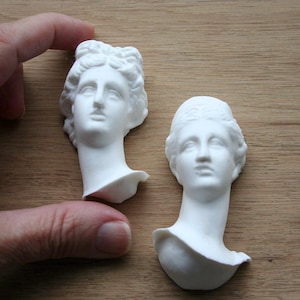 May include: Two white plaster busts of classical figures. One is held by a hand, the other rests on a wooden surface. The busts feature detailed facial features and stylized hair, with a smooth, matte finish.