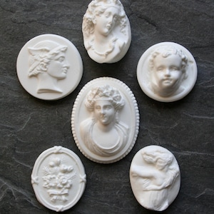 Peut inclure: Une collection de six pendentifs camées blancs de différentes formes ovales et rondes. Les camées présentent des motifs en relief classiques, notamment des portraits de femmes, des chérubins et des compositions florales. Les pendentifs sont exposés sur une surface gris foncé.