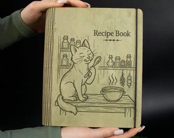 Puede incluir: Un libro de recetas de madera con una ilustración de un gato. La portada presenta las palabras "Recipe Book" sobre un dibujo de un gato sosteniendo una cuchara, con una olla de comida y estantes de frascos. El libro está abierto por manos.