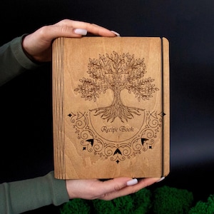 Può includere: Un libro di ricette in legno con un disegno dell'albero della vita inciso sulla copertina. Le parole "Recipe Book" sono sotto l'albero. Il libro è tenuto da mani su uno sfondo scuro.