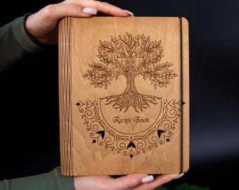 May include: A wooden recipe book with a tree of life design engraved on the cover. The words "Recipe Book" are below the tree. The book is held by hands against a dark background.