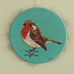 Op de afbeelding: Een roodborstje van stof is gestikt op een turquoise rond canvas met een witte rand. De vogel is bruin, rood en wit met een lange staart en een kleine snavel.