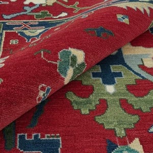 May include: Close-up of a rolled-up red rug with a detailed pattern. The rug features a central red field with a border of blue, green, and cream-colored designs. The intricate pattern is visible in the rolled section.
