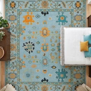 Könnte beinhalten: Draufsicht auf einen großen, rechteckigen Teppich mit hellblauem Hintergrund und aufwendigen Blumenmustern in Orange, Türkis und Gold. Der Teppich befindet sich in einem Schlafzimmer, teilweise unter einem Bett.