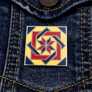 May include: A square-shaped enamel pin with a geometric design in red, blue, and yellow. The pin features a starburst pattern with a yellow background and a red and blue border.