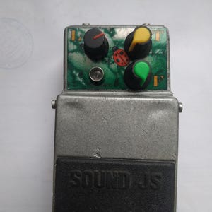 以下が含まれることがあります： SOUND JSと刻印された黒いフットスイッチを備えた銀色の金属製ギターエフェクトペダル。トップパネルは緑色のマーブル模様で、テントウムシのイラスト、3つのコントロールノブ、金属製のインジケーターライトが配置されています。