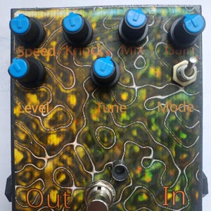 Puede incluir: Un pedal de efectos para guitarra rectangular con un patrón jaspeado en negro y amarillo. Cuenta con cinco perillas negras con tapas azules, un interruptor de palanca metálico y un interruptor de pie. Las etiquetas Speed, Knock, Mix, Gain, Level, Tune, Mode, Out e In están impresas en naranja.