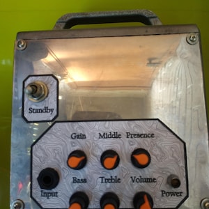 May include: A metal guitar amplifier head featuring a clear front panel with a topographic map pattern. It includes a Standby toggle switch, an Input jack, and six black knobs with orange indicators labeled Gain, Middle, Presence, Bass, Treble, and Volume.