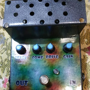 Puede incluir: Un pedal de efectos de guitarra personalizado con un diseño de camuflaje. Cuenta con cuatro perillas de control etiquetadas como "LEVEL", "COMP", "DRIVE" y "GAIN", además de conectores "OUT" e "IN". La parte superior está cubierta con una placa de metal perforada.