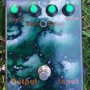 May include: A rectangular green and black effects pedal with four green knobs and a silver foot switch. The pedal has text that reads "Level", "Tone", "Comp", "Gain", "Output", and "Input". The pedal is on a grassy surface.