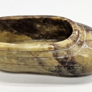 May include: A carved stone ashtray or small bowl. The oval-shaped container is made of tan and brown stone with a smooth, polished finish. The bowl has a rounded rim and a carved design on the side. The interior is a darker brown color.