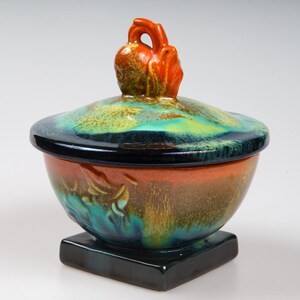 May include: A ceramic bowl with a lid, featuring a vibrant orange, green, and blue glaze. The lid has a unique, stylized handle in the shape of a bird.