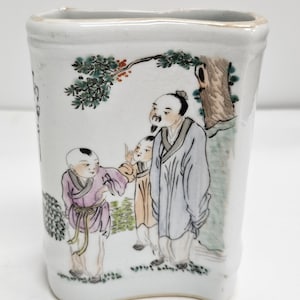 Puede incluir: Un jarrón de cerámica blanca con forma rectangular y base ligeramente curvada. El jarrón presenta una escena pintada a mano de tres figuras bajo un árbol, con pinceladas detalladas y colores vibrantes. Las figuras visten atuendos tradicionales.