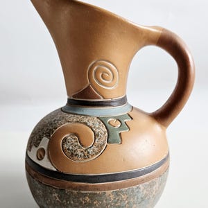 Puede incluir: Una jarra de cerámica con cuerpo marrón y base redondeada. La jarra presenta un asa curva y un pico. Patrones decorativos en espiral y geométricos en marrón, verde y azul adornan el cuerpo. La base tiene un aspecto moteado y texturizado.