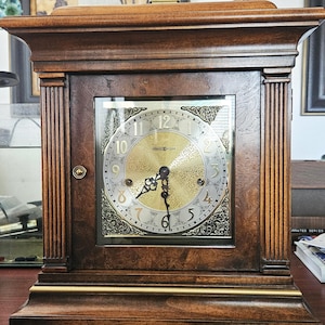 May include: A wooden Howard Miller mantel clock with a gold-toned face and Roman numerals. The clock has a rectangular shape with decorative columns on the sides and a brass handle on top. The clock face is visible through a glass panel.