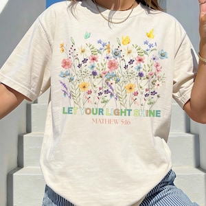 Puede incluir: Camiseta color crema con un diseño floral colorido y mariposas. El texto "LET YOUR LIGHT SHINE" y "MATTHEW 5:16" está impreso debajo de las flores. La persona lleva pantalones a rayas azules y blancas.