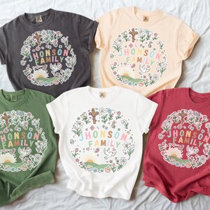 Puede incluir: Cinco camisetas de manga corta en varios colores: carbón, melocotón, verde, blanco y rojo. Cada camiseta presenta un diseño de corona floral con el texto "JHONSON FAMILY" en letras coloridas. El diseño incluye una cruz, una paloma y un amanecer.