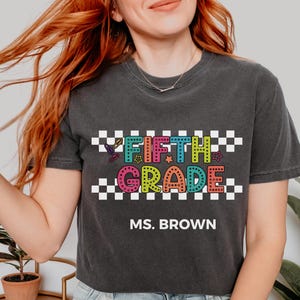 May include: A charcoal gray t-shirt featuring a graphic with a black and white checkered pattern background. The text reads FIFTH GRADE in colorful, dotted, block letters with small star and paperclip icons. Below, the text reads MS. BROWN.