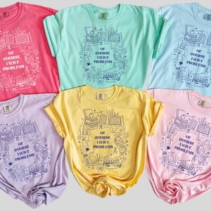 Puede incluir: Una colección de seis camisetas de manga corta y cuello redondo en rosa, verde menta, azul claro, lavanda, amarillo y rosa claro. Cada camiseta presenta un estampado gráfico azul oscuro de material escolar y el texto OF COURSE I HAVE PROBLEMS.