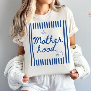 Puede incluir: Camiseta color crema con la palabra "Motherhood" en escritura azul dentro de un marco rectangular azul. El diseño incluye líneas verticales azules e ilustraciones de estrellas. Una prenda casual para el día a día.