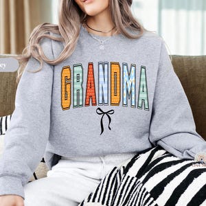 Puede incluir: Una sudadera gris deportivo de cuello redondo con la palabra GRANDMA impresa en letras grandes y multicolores con varios patrones que incluyen lunares, cuadros y flores. Un pequeño lazo negro está dibujado debajo del texto.