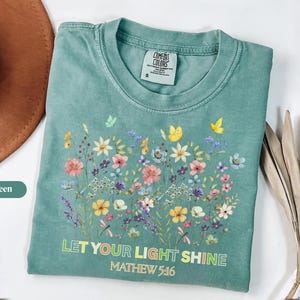 Puede incluir: Camiseta verde claro con un diseño floral con flores y mariposas de colores. El texto "LET YOUR LIGHT SHINE" y "MATTHEW 5:16" está impreso en la parte delantera. La camiseta es de la marca Comfort Colors.