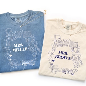 Puede incluir: Una camiseta azul con dibujos de líneas blancas y una camiseta crema con dibujos de líneas azul oscuro. Ambas presentan un diseño circular de material escolar, un autobús y libros que rodean el texto MRS. MILLER y MRS. BROWN respectivamente.