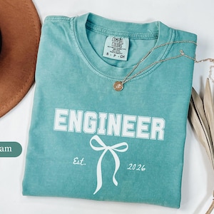 Puede incluir: Camiseta verde espuma con la palabra "ENGINEER" en letras blancas. Debajo, un lazo blanco y el texto "Est. 2026". Un collar dorado y un sombrero marrón también están en la imagen.