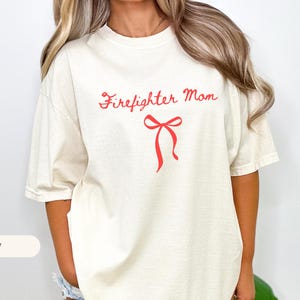 Puede incluir: Camiseta de color marfil con el texto "Firefighter Mom" en rojo, acompañada de un lazo rojo. La camiseta tiene mangas cortas y un corte holgado. El color de la camiseta es marfil.