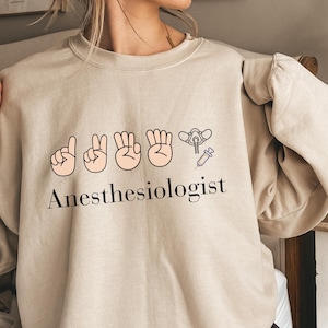 Puede incluir: Una sudadera beige de cuello redondo con un estampado gráfico de cuatro manos mostrando dedos en secuencia, una máscara de anestesia, una jeringuilla y el texto Anesthesiologist en letras negras en el centro del pecho.