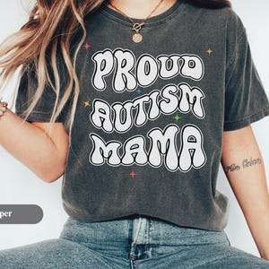 Puede incluir: Una camiseta gris oscuro con las palabras "Proud Autism Mama" en blanco, con una fuente de estilo burbuja. La camiseta tiene un aspecto vintage y descolorido y presenta acentos de estrellas de colores. La palabra "Pepper" está en un rectángulo gris en la parte inferior.