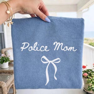 以下が含まれることがあります： 白い筆記体で「Police Mom」と白いリボン模様が描かれた青い布製品。手で持っており、背景にはベランダと海の景色が写っています。