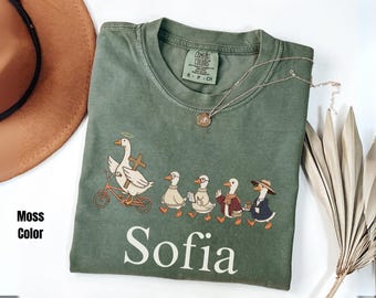 May include: Moss green t-shirt with a whimsical goose design. The graphic features a goose riding a bicycle, followed by other geese in various outfits. The name "Sofia" is printed in large white letters.