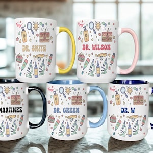 Puede incluir: Tazas de cerámica blanca con asas e interiores de colores. Cada taza presenta un nombre, como "Dr. Smith" o "Dr. Wilson", y está decorada con ilustraciones de temática científica, incluyendo microscopios y soles.