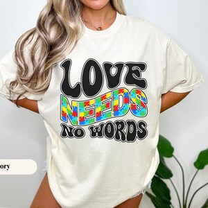 Puede incluir: Camiseta de color marfil con la frase "LOVE NEEDS NO WORDS" en negro, con una fuente retro. La palabra "NEEDS" está rellena con un patrón de rompecabezas colorido. La camiseta es de estilo casual y unisex.