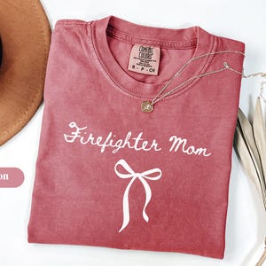 Puede incluir: Camiseta de color carmesí con las palabras "Firefighter Mom" en escritura blanca y un lazo blanco. La camiseta tiene cuello redondo y mangas cortas. Un collar dorado con un colgante está sobre la camiseta.