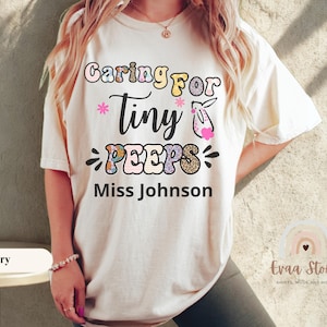Może przedstawiać: Kremowa koszulka z napisem "Caring for Tiny Peeps" w kolorowych literach, z motywem królika i imieniem "Miss Johnson". Koszulka ma luźny, wygodny krój.