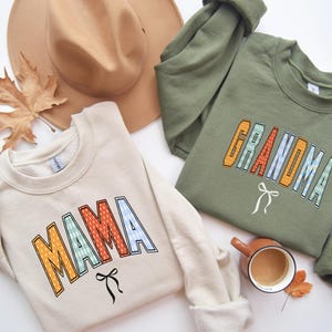 Könnte beinhalten: Ein cremefarbenes Sweatshirt mit dem Wort MAMA in orange, grün und blau gemusterten Buchstaben mit einer schwarzen Schleife und ein olivgrünes Sweatshirt mit dem Wort GRANDMA in ähnlichen Buchstaben mit einer weißen Schleife.