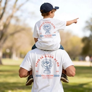 Puede incluir: Camisetas blancas con gráficos a juego. Las camisetas presentan un diseño de trofeo con el texto "SPORTS KIDS CLUB" y "SPORTS DADS CLUB". También es visible el texto "EST. 2026" y "STILL COACHING JUST TIRED".