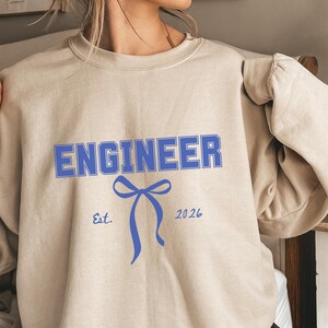 Puede incluir: Sudadera beige con la palabra "ENGINEER" en letras azules. Debajo, un diseño de lazo azul y el texto "Est. 2026". La sudadera tiene cuello redondo y mangas largas.